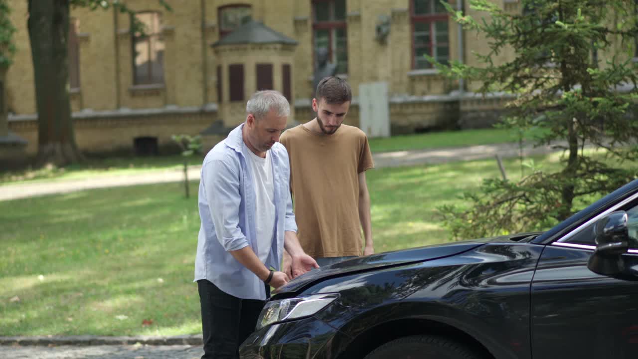 自信的中年男子帮助压力的家伙与破车在夏天的道路上。父亲修理被儿子弄坏的汽车的侧视图肖像。交通事故的概念。视频素材