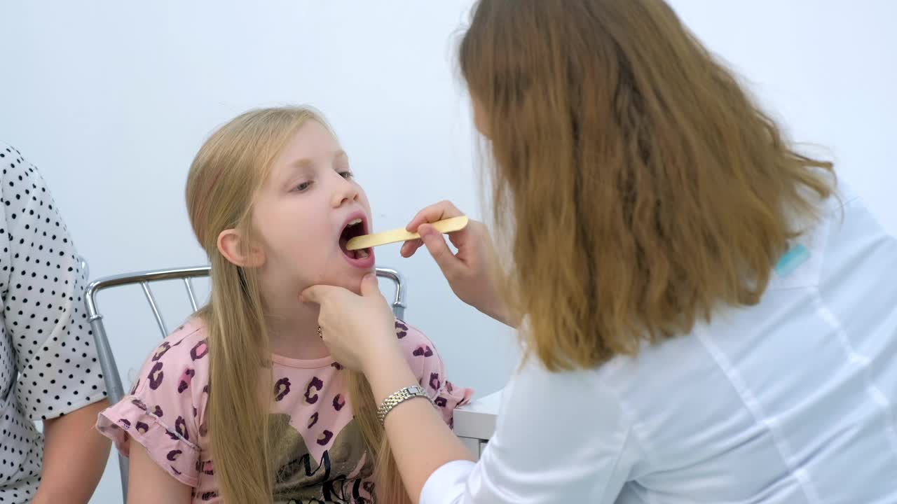 儿科医生妇女用刮刀和提灯看小女孩喉咙痛。视频素材