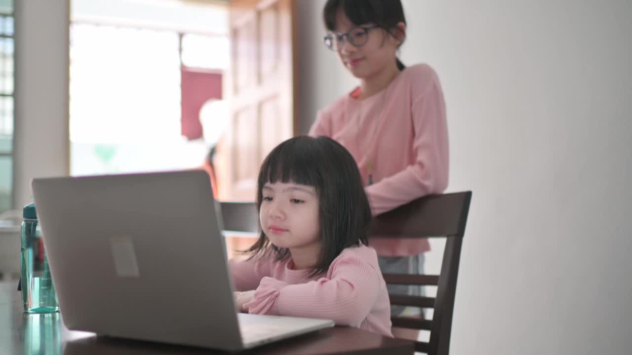 亚裔中国女孩在她姐姐的帮助下在厨房餐桌上参加网络课程视频素材