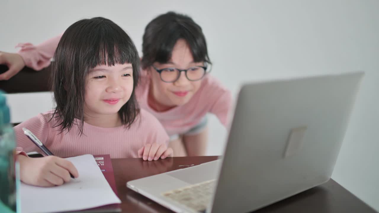 亚裔中国女孩在她姐姐的帮助下在厨房餐桌上参加网络课程视频素材