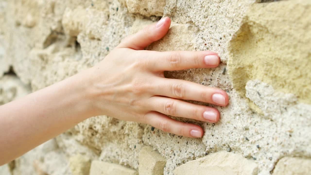 一个女人的手缓慢地靠在古老的石墙上。女性的手触摸岩石的粗糙表面视频素材