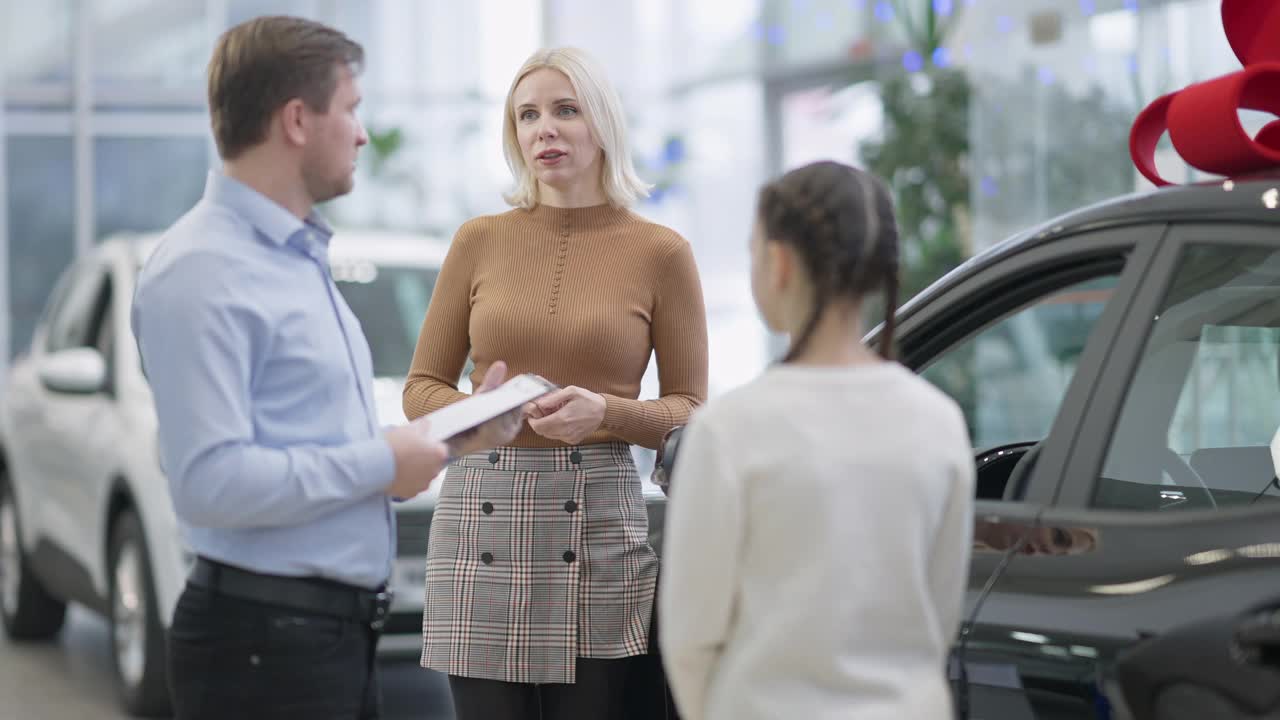 肖像自信的金发女人与汽车经销商与女儿选择新车交谈。苗条迷人的白人母亲咨询雇员在汽车经销商。奢侈和财富的概念。视频素材