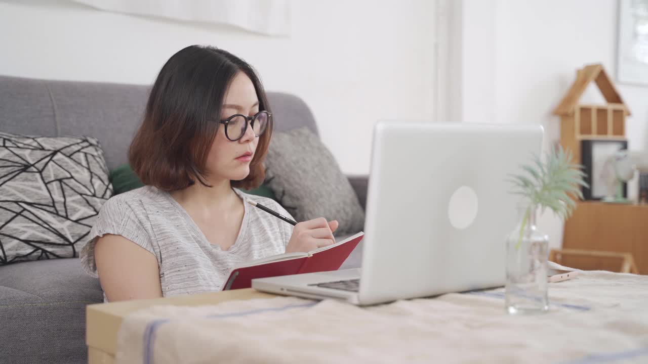 下午在家远程办公，集中亚洲妇女坐在客厅的地板办公桌上，打字电脑笔记本键盘和在记事本上写笔记。在网上收集信息。视频素材