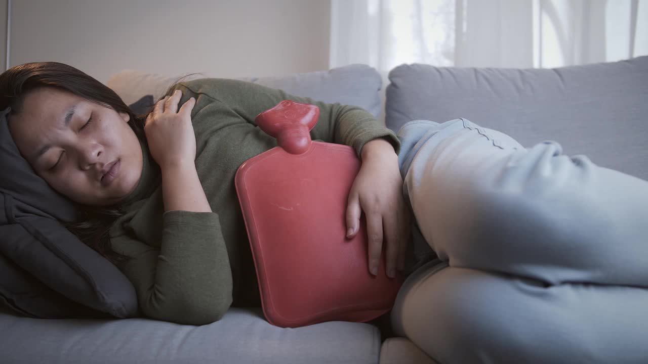 一名痛经严重的妇女坐在客厅沙发上，用热水袋敷在肚子上缓解疼痛，感觉好多了视频素材