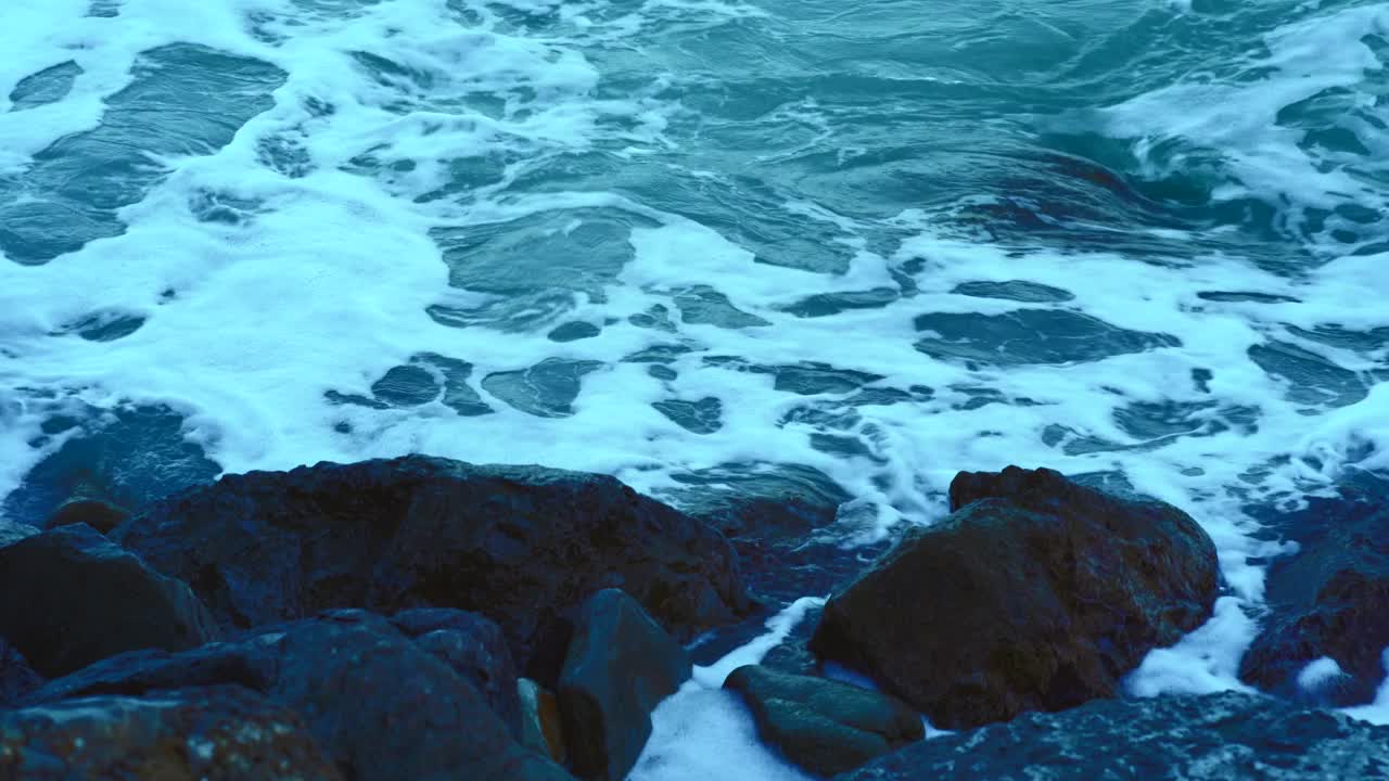 海水的浪花溅起泡沫溅向海岸的岩石，呈蓝色调视频素材