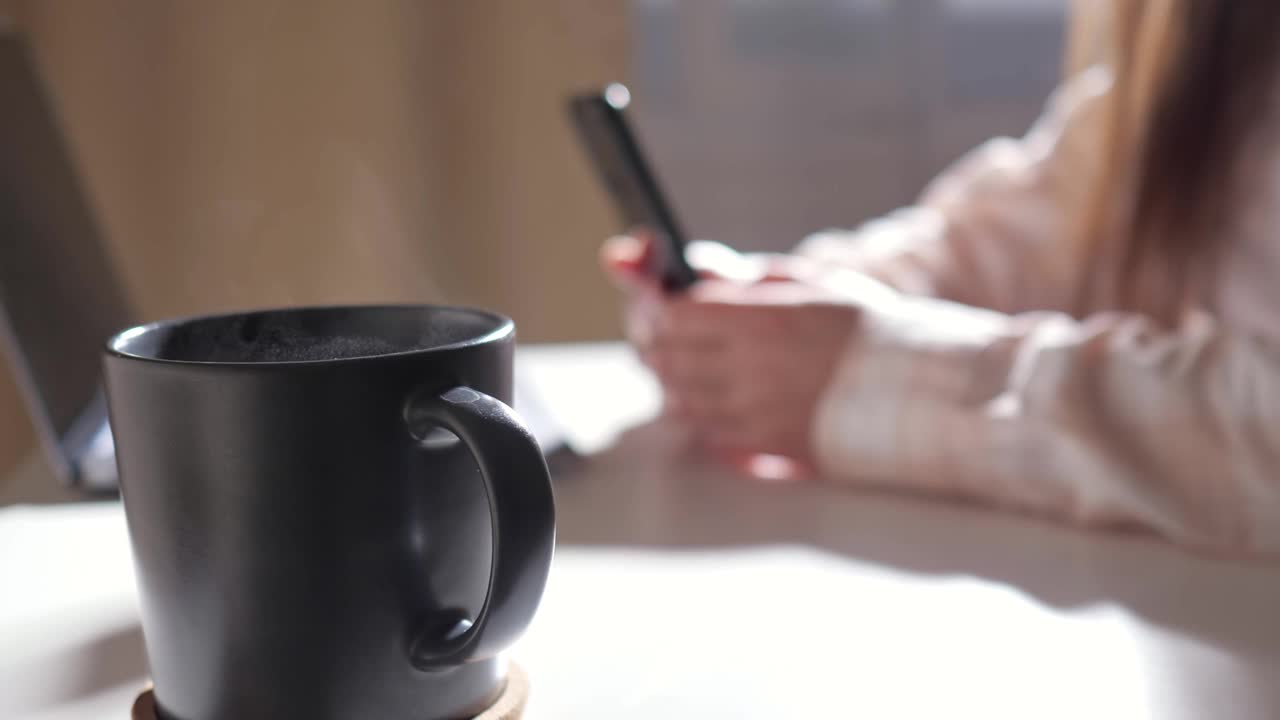 特写的杯子与热饮料在桌子上的背景下，一个不认识的女人与手机和笔记本电脑视频素材