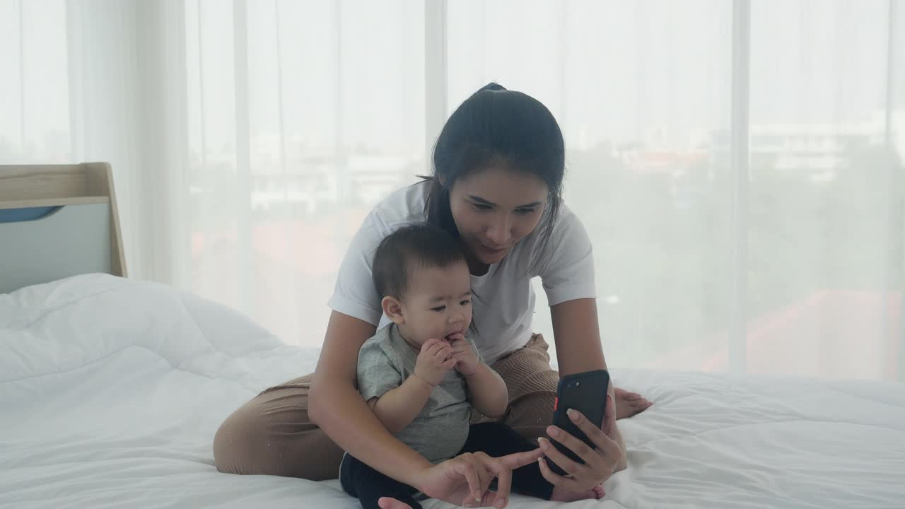 年轻的亚洲妈妈和小女婴或新生儿在卧室的床上用智能手机自拍，幸福的妈妈和女儿在家里用手机视频通话，两人，家庭和沟通的概念。视频素材