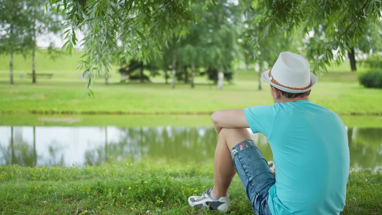 一个戴着帽子的年轻人坐在城市公园的岸边，背景视频素材