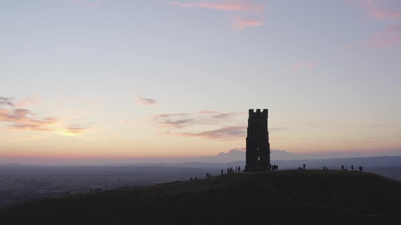 在英国萨默塞特，一架环绕格拉斯顿伯里山的夜航飞机和游客们在五彩缤纷的天空下的剪影视频素材