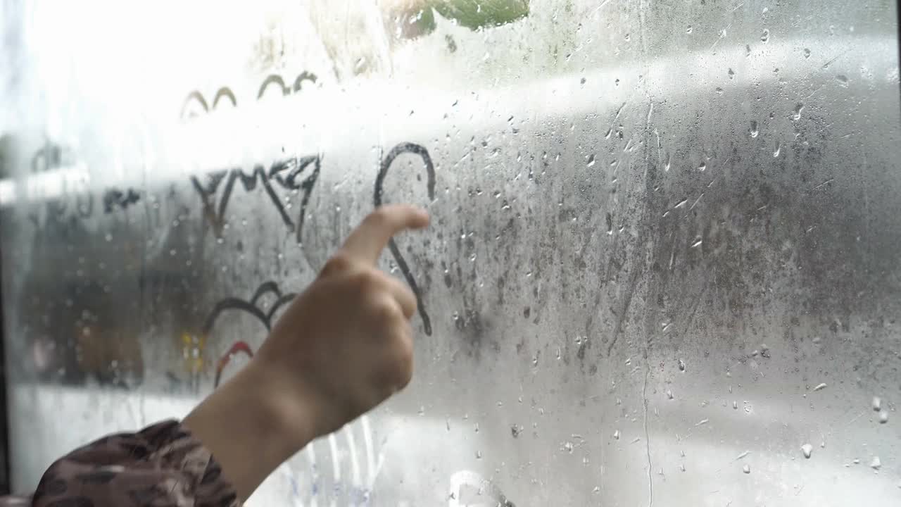 一个孩子用他的手指在一个阴沉的雨天穿过城市的巴士雾蒙蒙的玻璃上画了一颗心。汽车在窗外行驶。这个概念。不知名的。特写镜头。4 k。视频素材