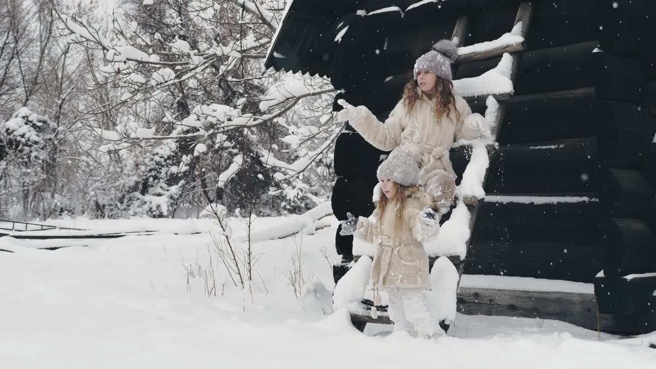 冬天的肖像。可爱、漂亮的小女孩们，穿着暖和的冬装，浑身沾满了雪花，在户外玩耍，靠近老木屋。雪天快乐时光视频素材