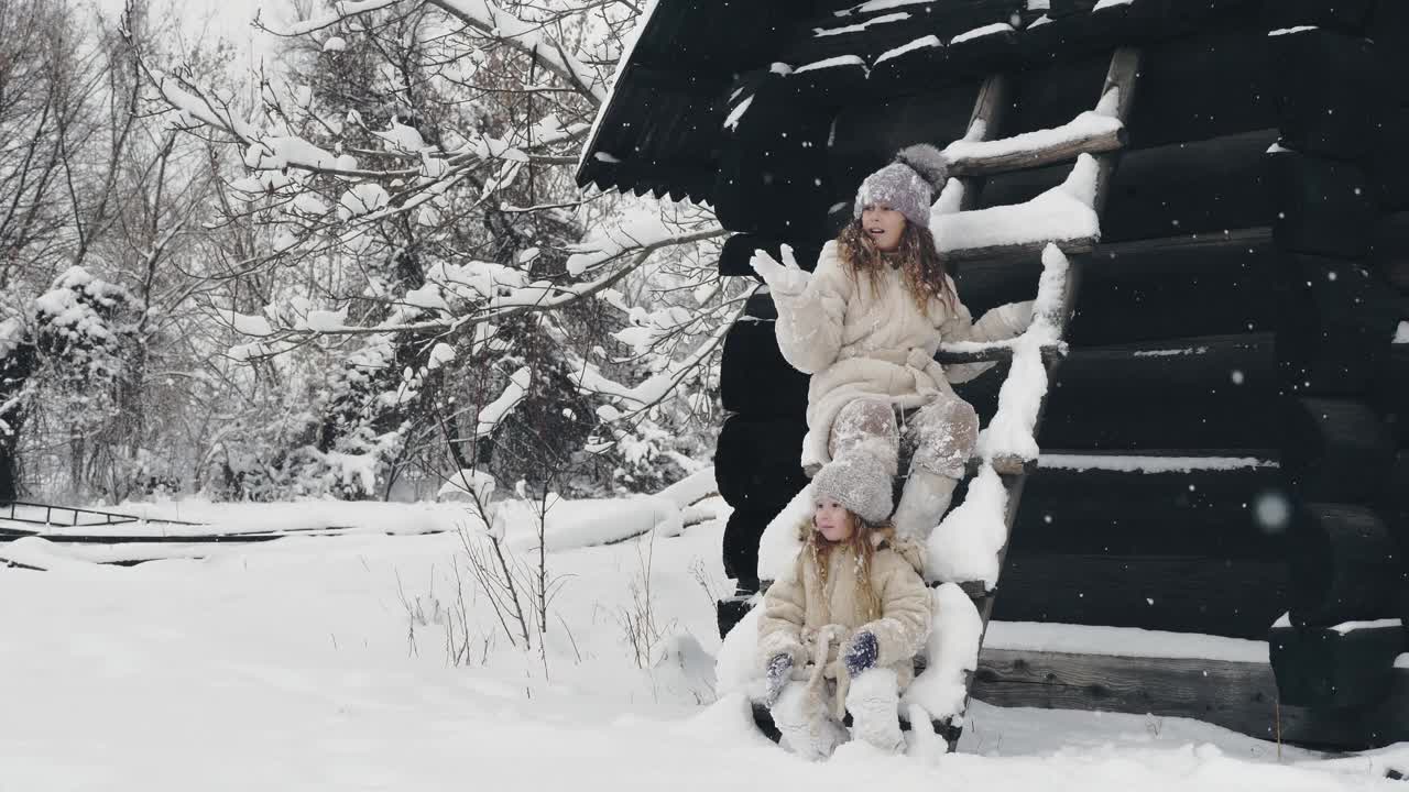 冬天的肖像。可爱、漂亮的小女孩们，穿着暖和的冬装，浑身沾满了雪花，在户外玩耍，靠近老木屋。雪天快乐时光视频素材