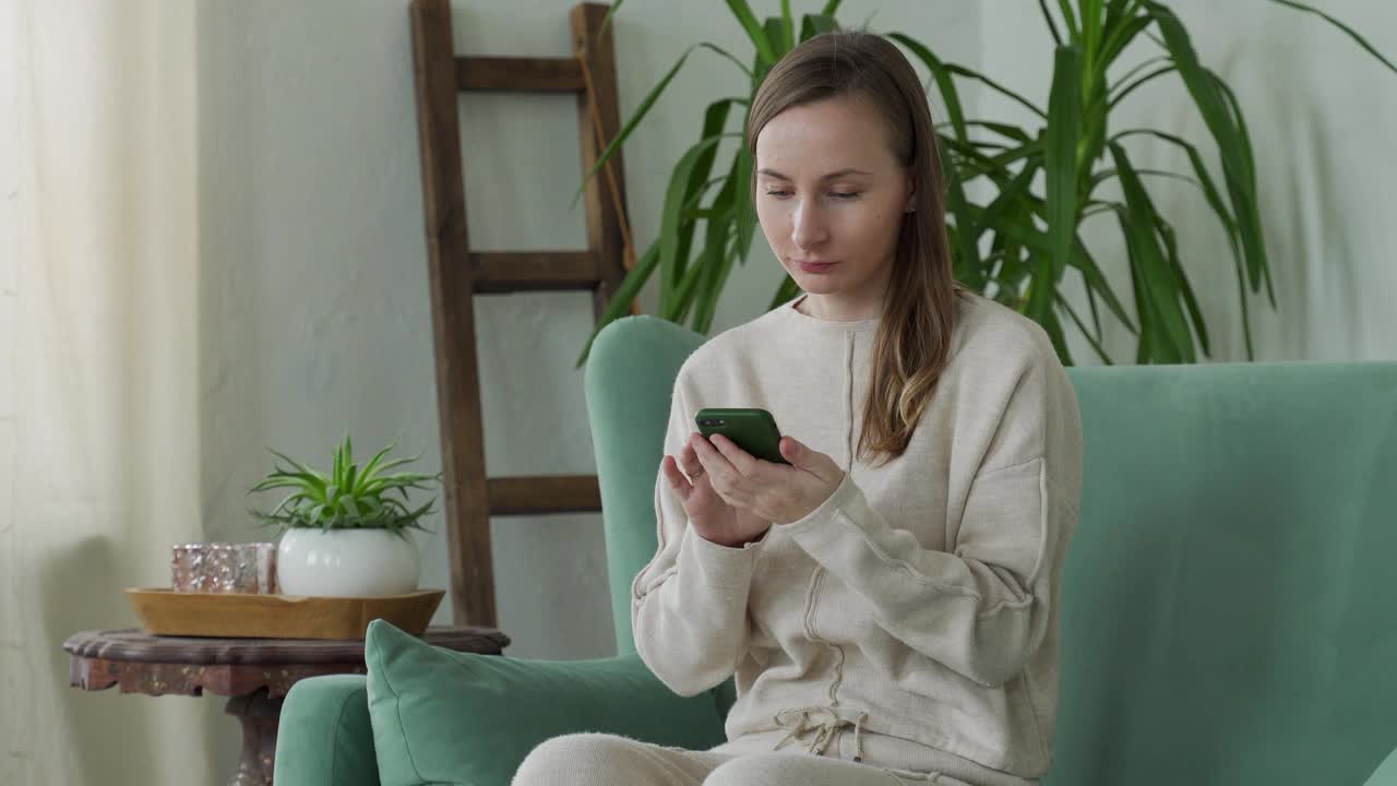 年轻女子用手机，庆祝网上彩票中奖，阅读好消息，坐在家里的沙发上视频素材