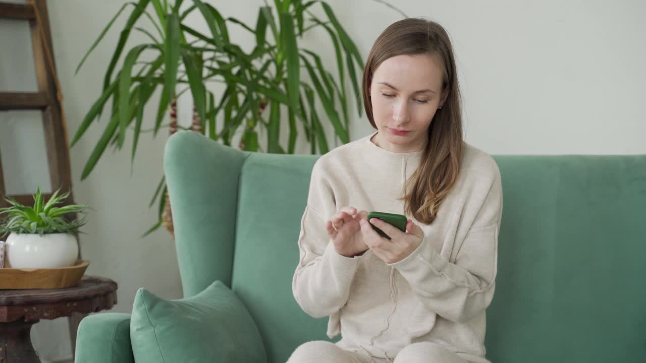 年轻女子用手机，庆祝网上彩票中奖，阅读好消息，坐在家里的沙发上视频素材