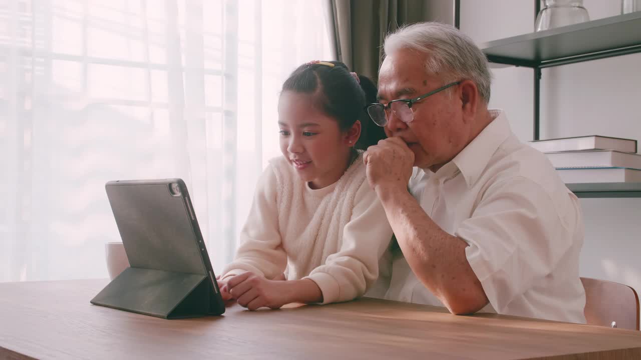 一位亚洲祖父和孙女在客厅里共度时光。快乐的老人和小女孩使用平板电脑在家里的桌子上。视频素材