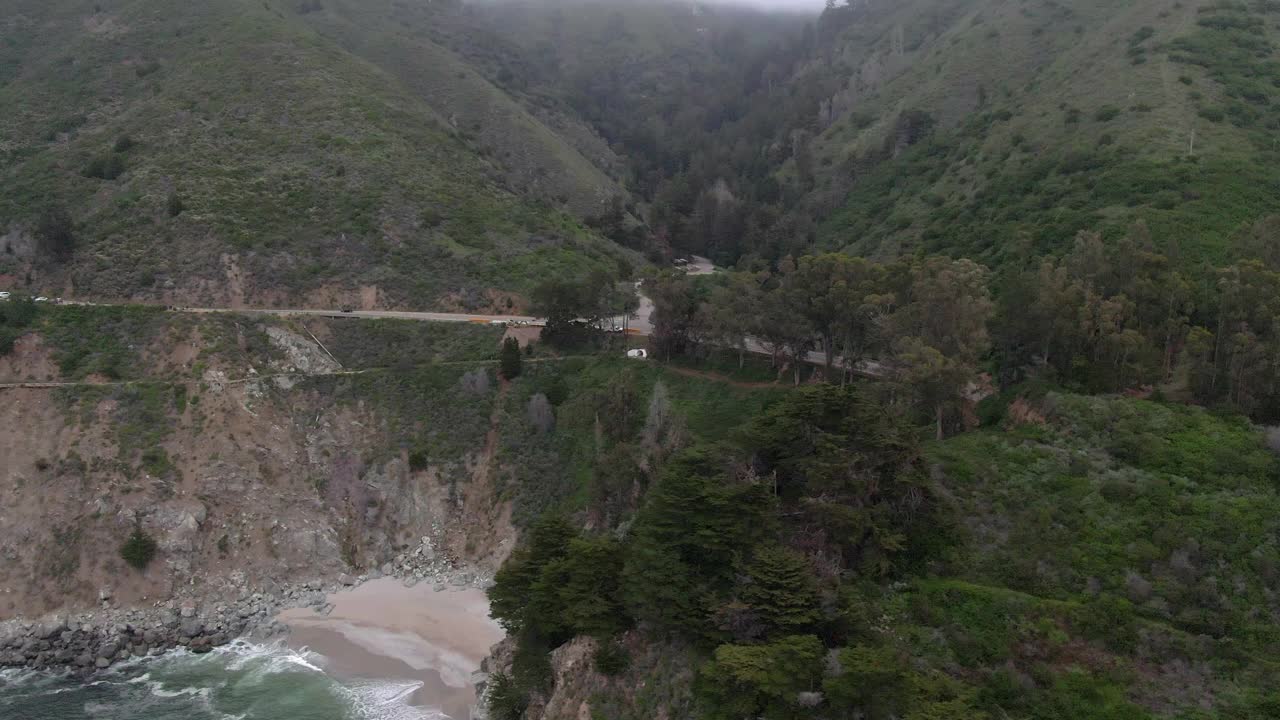 空中拍摄的车辆在道路上的树木由麦克威瀑布，无人机飞过海浪溅在海滩对山脉-大苏尔，加利福尼亚视频素材