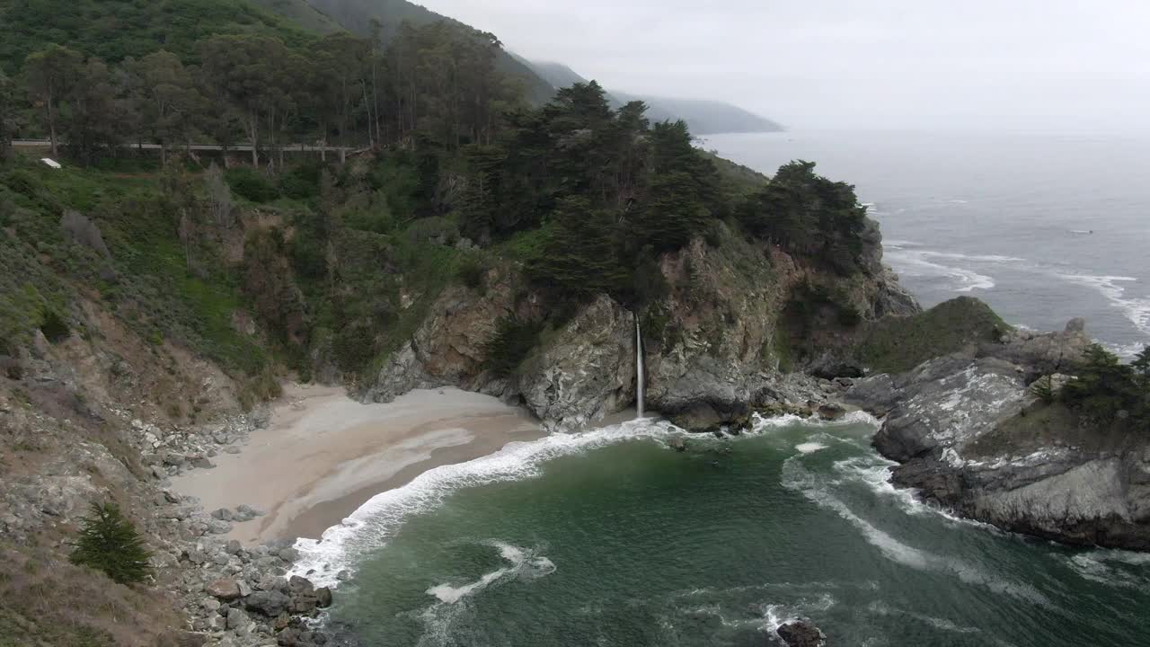 空中拍摄的麦克威瀑布从岩石悬崖在海滩，无人机下降在海浪拍打海岸-大苏尔，加利福尼亚视频素材