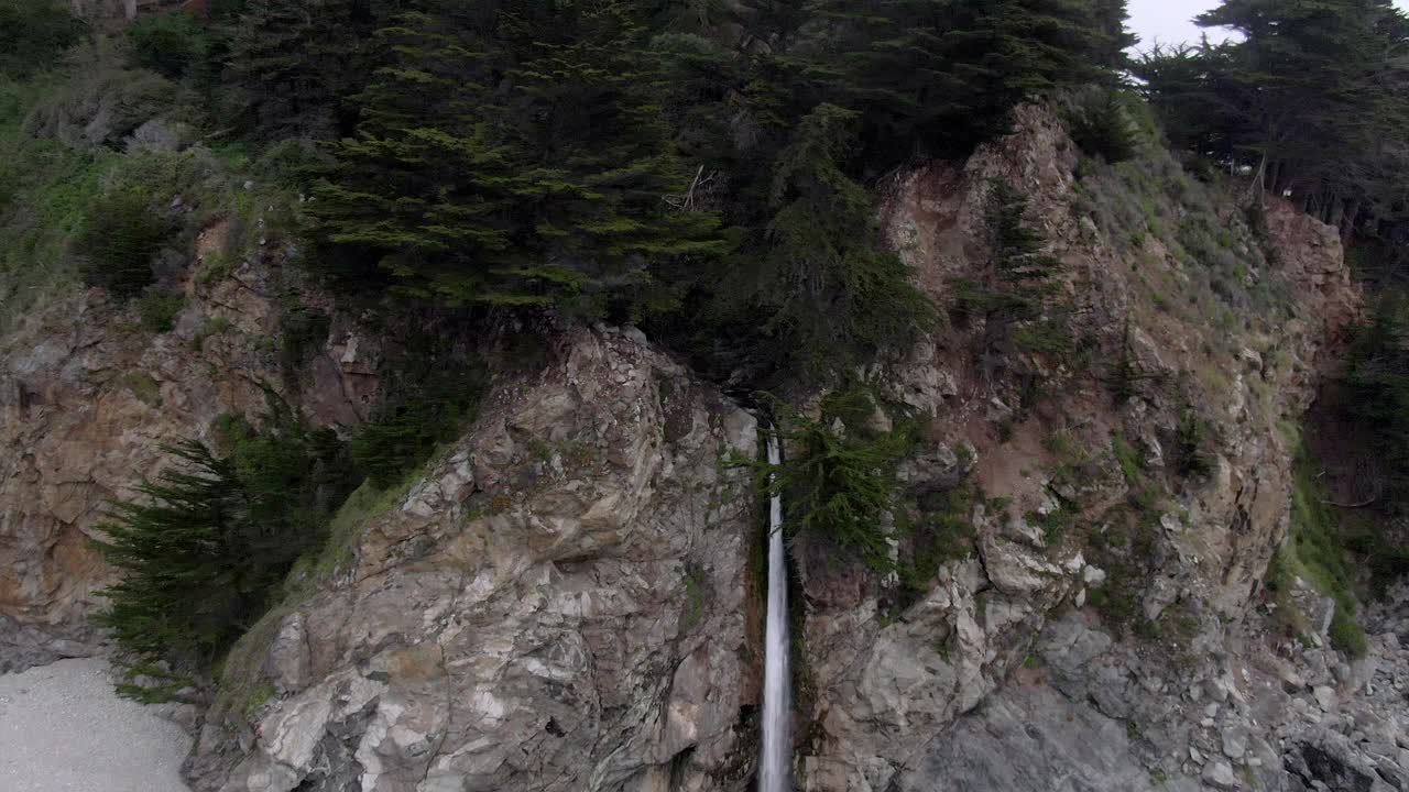 航拍美丽的麦克威瀑布从岩石悬崖在海滩上，无人机向后飞行在海浪拍打海岸-大苏尔，加利福尼亚视频素材