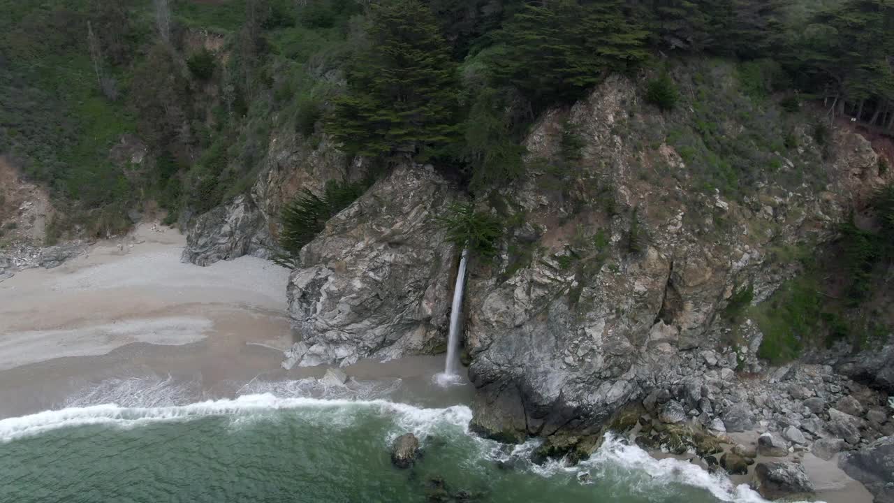 空中摇摄麦克威瀑布从岩石悬崖上的海岸，无人机飞过浪花溅在海滩-大苏尔，加利福尼亚视频素材