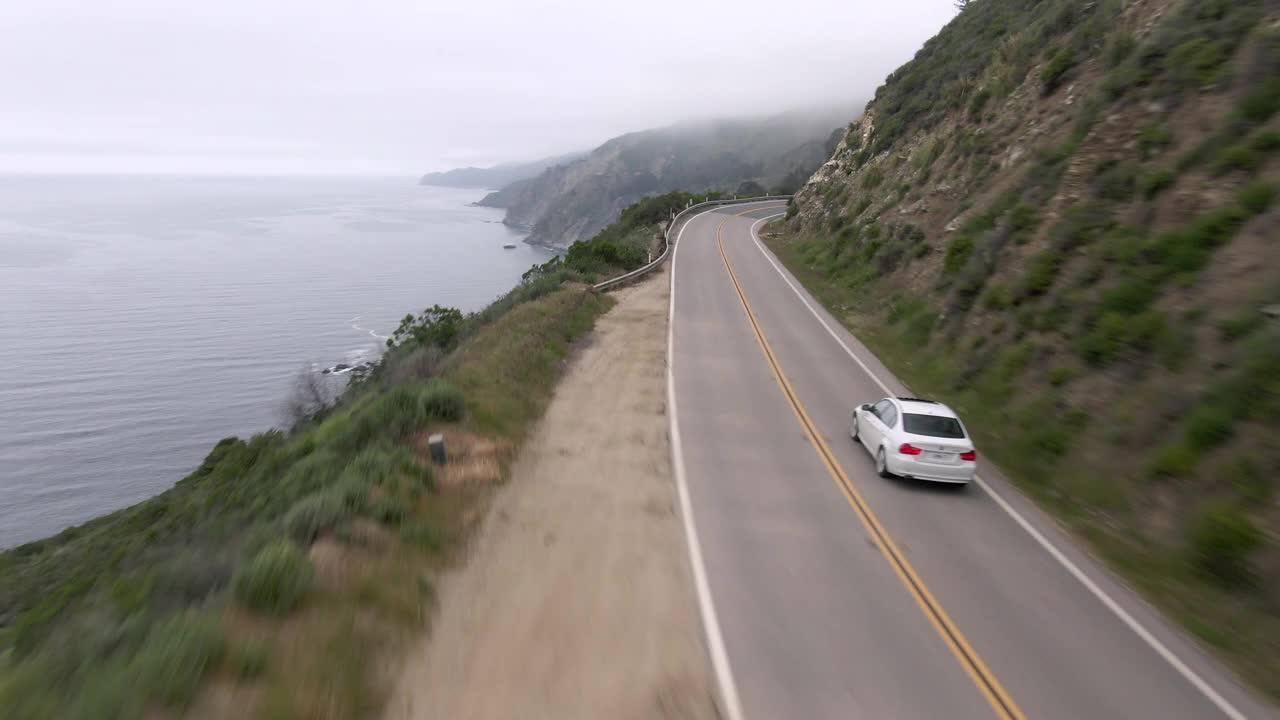 空中拍摄的白色汽车在蜿蜒的海岸公路上的山，无人机向前飞行海岸线对天空-大苏尔，加利福尼亚视频素材