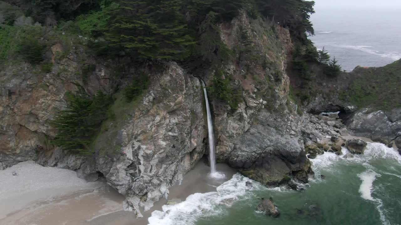 航拍拍摄著名的麦克威瀑布从岩石悬崖上的海岸，无人机飞过浪花溅在海滩-大苏尔，加利福尼亚视频素材