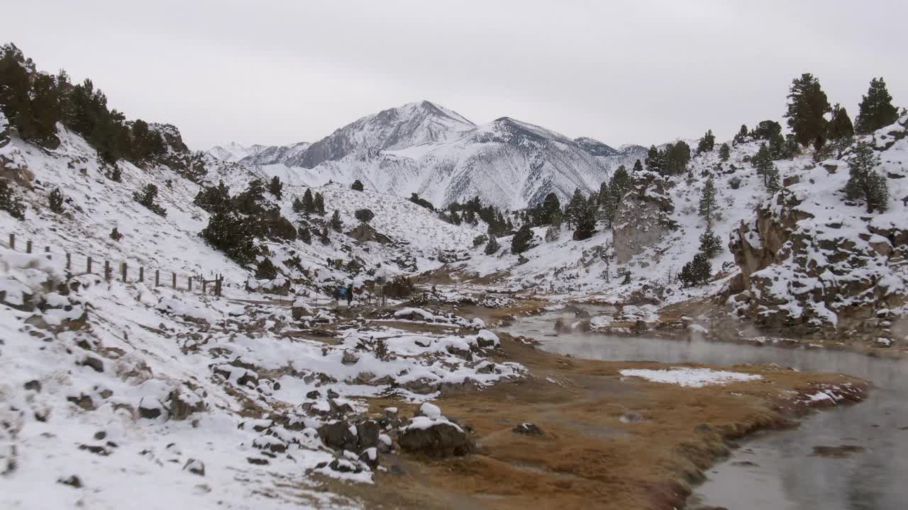 航空拍摄的游客在热溪地质地点在雪中，无人机向前飞行在溪附近的雪顶山脉对天空-猛犸湖，加利福尼亚视频素材