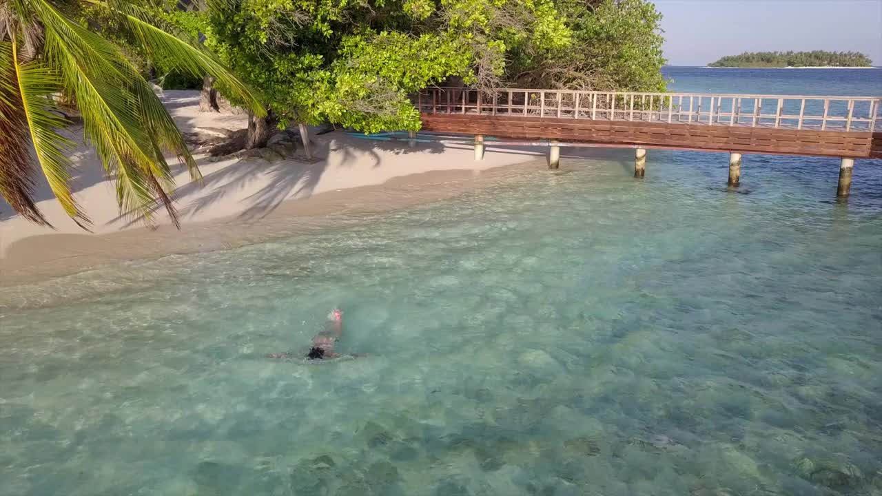 航拍的女人游泳在绿松石海，无人机从水上小屋向后上升对天空在阳光明媚的一天-南马莱环礁，马尔代夫视频素材