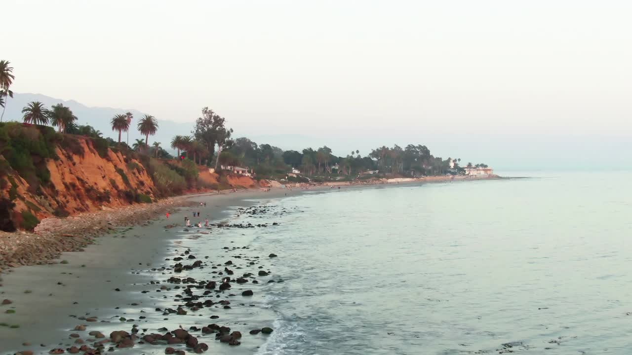 空中拍摄的人在蝴蝶海滩对天空，无人机向后上升在海浪溅在海岸在日落-圣巴巴拉，加利福尼亚视频素材