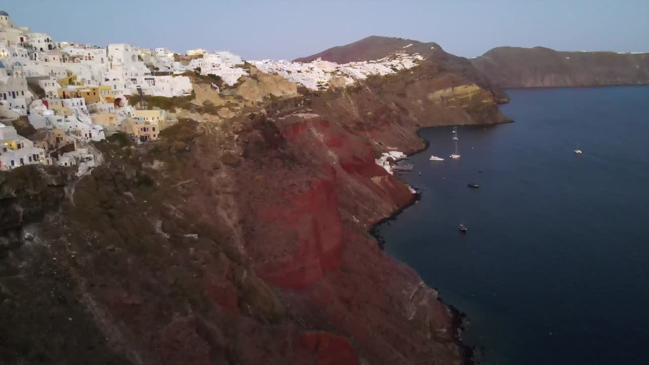 航拍在海岸线上的船在山脉上的城市，无人机在日落时对着天空在海上上升-圣托里尼，希腊视频素材