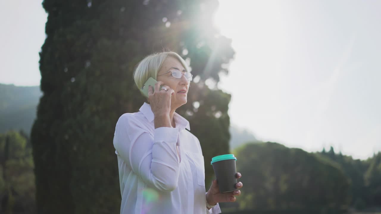 一位年长的女士在城市公园里用手机跟人聊天喝着咖啡视频素材