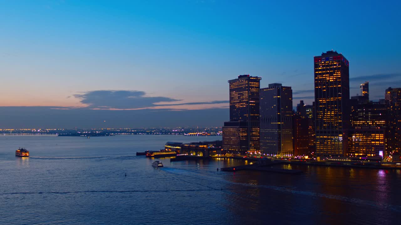 曼哈顿下城的鸟瞰图横跨东河照亮了夜晚，与新泽西海岸在地平线上的远景。无人机视频与向后和平移摄像机运动。视频素材