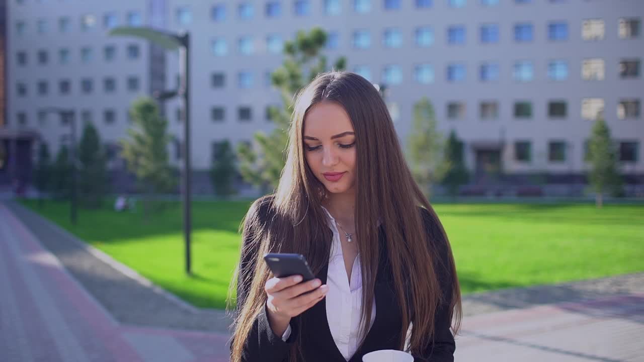 年轻的职业女商人或女学生，走路时使用手机、智能手机、发短信和喝咖啡的女性视频素材