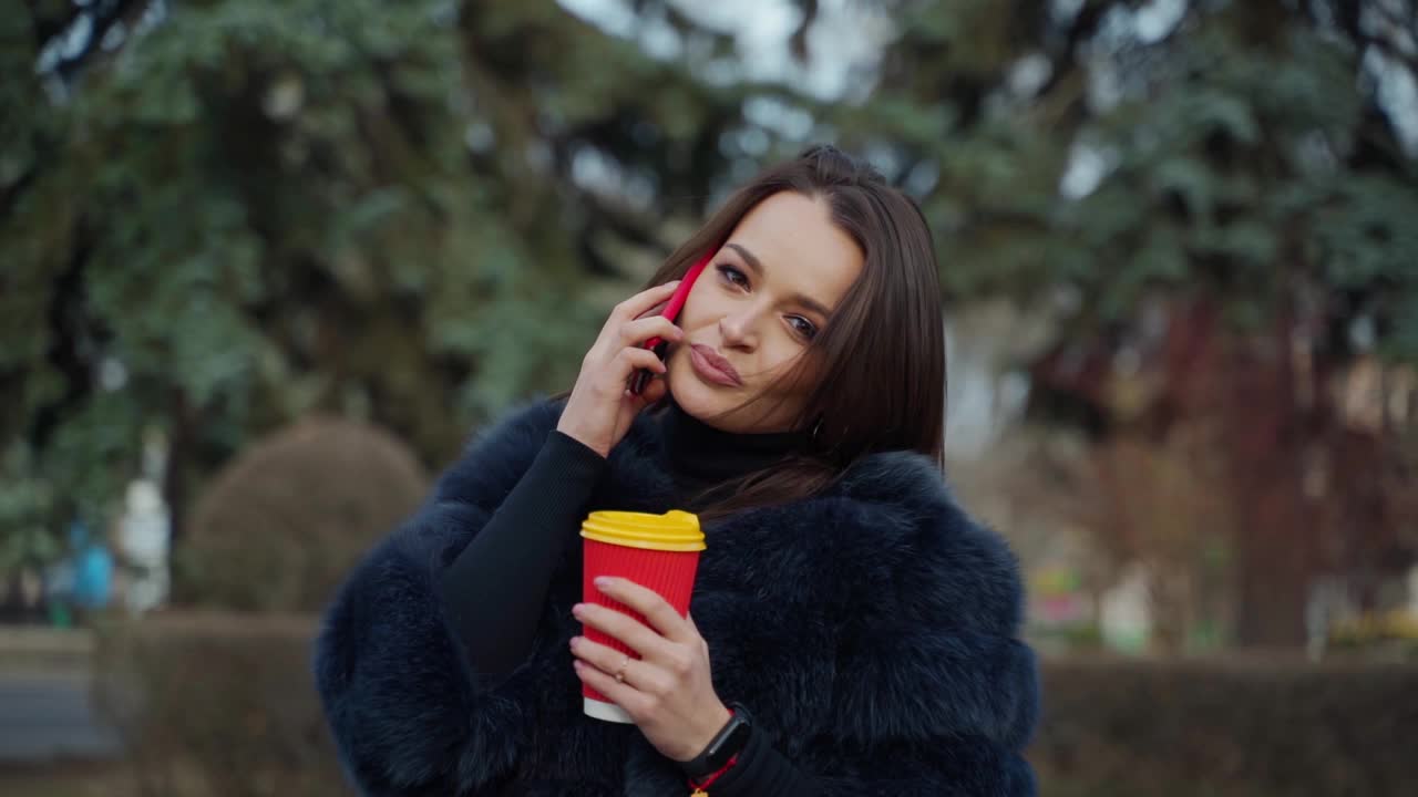 穿着裘皮大衣的迷人年轻女孩。漂亮的女人穿着时髦的外套，在公园里打电话，拿着咖啡杯。视频素材