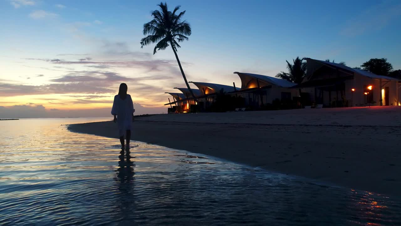 度假旅游剪影快乐的模特女孩穿着美丽的连衣裙走在海滩浪漫的度假酒店的夜晚。日落时分的热带岛屿豪华海滩别墅。造型在马尔代夫视频下载