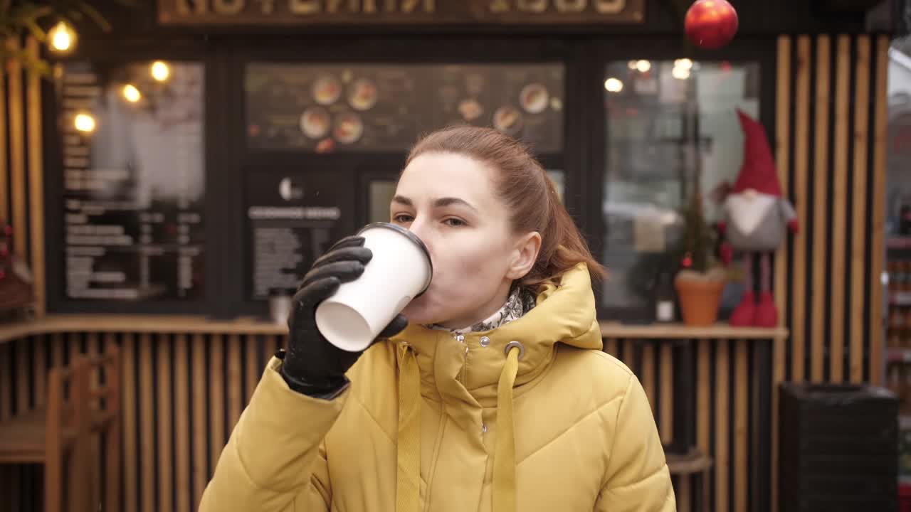 一个年轻的女人在街上的咖啡馆喝咖啡，在工作日之前的寒冷的早晨，享受早晨的咖啡，阁楼咖啡馆与外卖咖啡，慢镜头视频素材