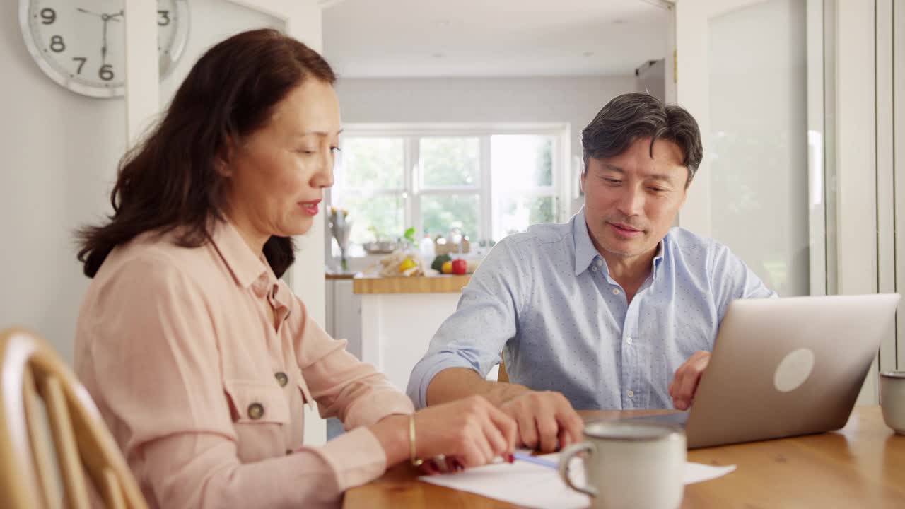 成熟的亚洲夫妇坐在家里的桌子旁，用笔记本电脑处理家庭账单和财务——慢镜头拍摄视频素材