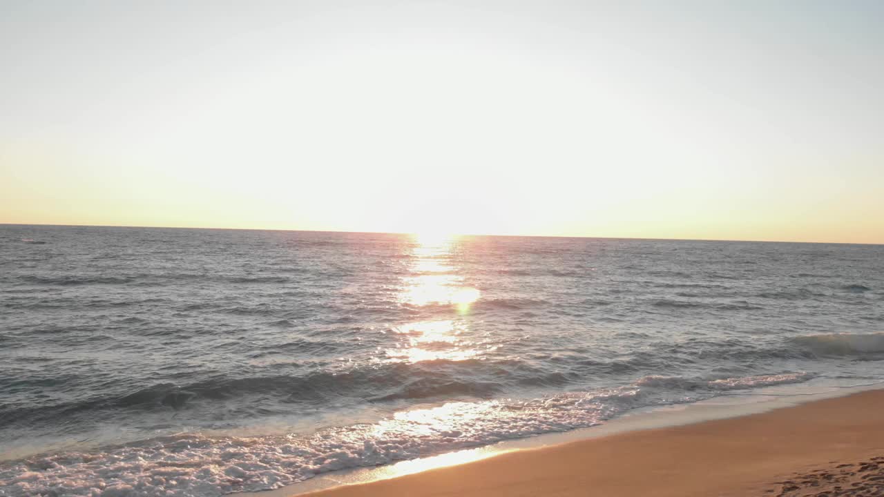 日落海滩。海浪拍打着沙滩，溅起水花。海面上红色的夕阳，太阳触地。夏季日落海景，阳光反射水面视频素材