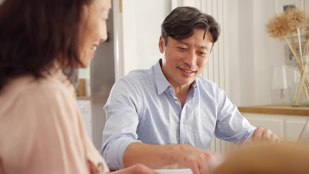 成熟的亚洲夫妇坐在家里的桌子旁，用笔记本电脑处理家庭账单和财务——慢镜头拍摄视频素材