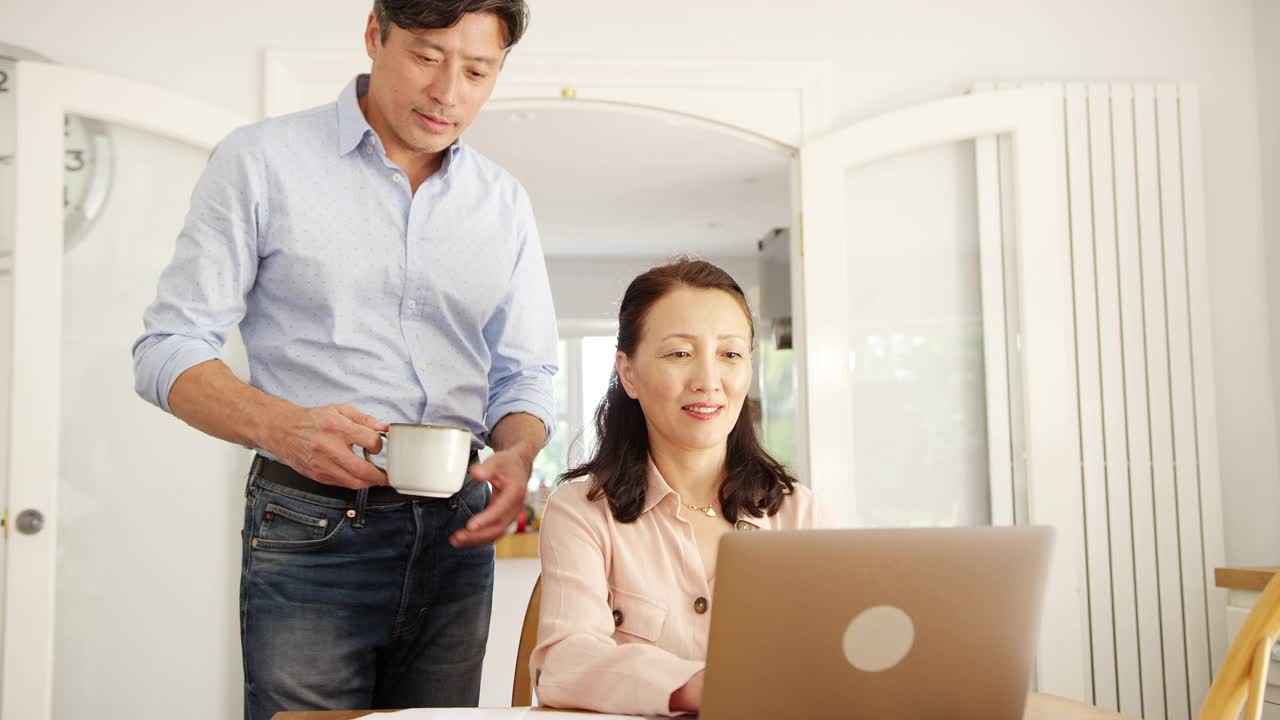 成熟的亚洲男人在家给妻子端咖啡，他们一起在笔记本电脑上查看家庭账单——慢镜头拍摄视频素材