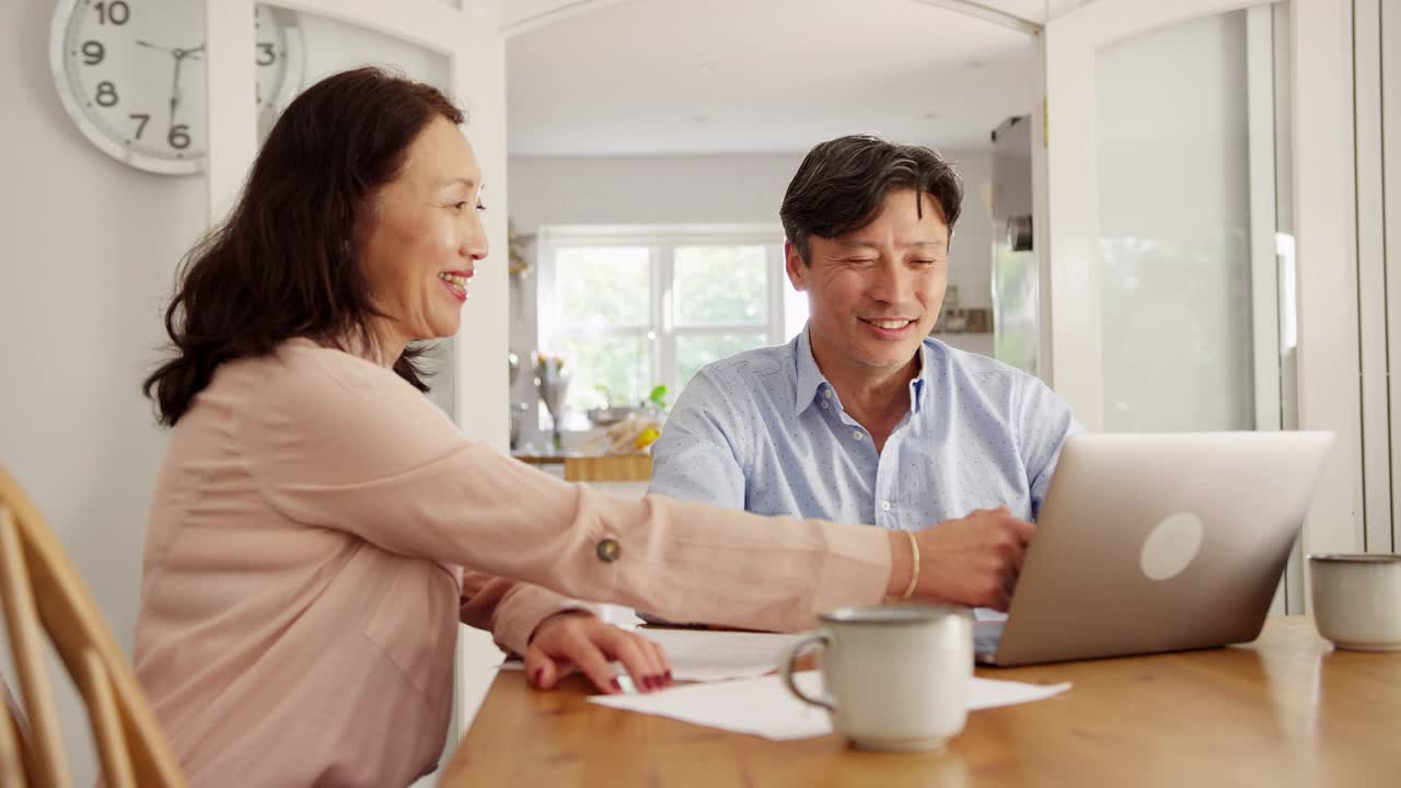 成熟的亚洲夫妇坐在家里的桌子旁，用笔记本电脑处理家庭账单和财务——慢镜头拍摄视频素材