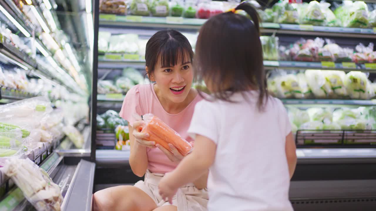 年轻的亚洲美丽的母亲拿着购物篮和她的孩子走在超市。她从架子上挑选沙拉蔬菜，然后给她的小孩吃。健康的购物。视频素材