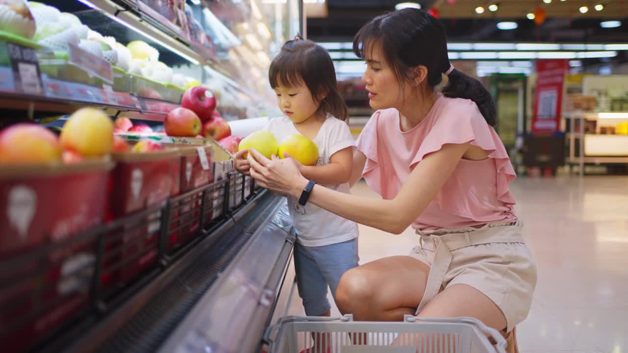 年轻的亚洲美丽的母亲拿着购物篮和她的孩子走在超市。她正在挑选红苹果水果，从架子上拿起，一起闻。健康的购物。视频素材