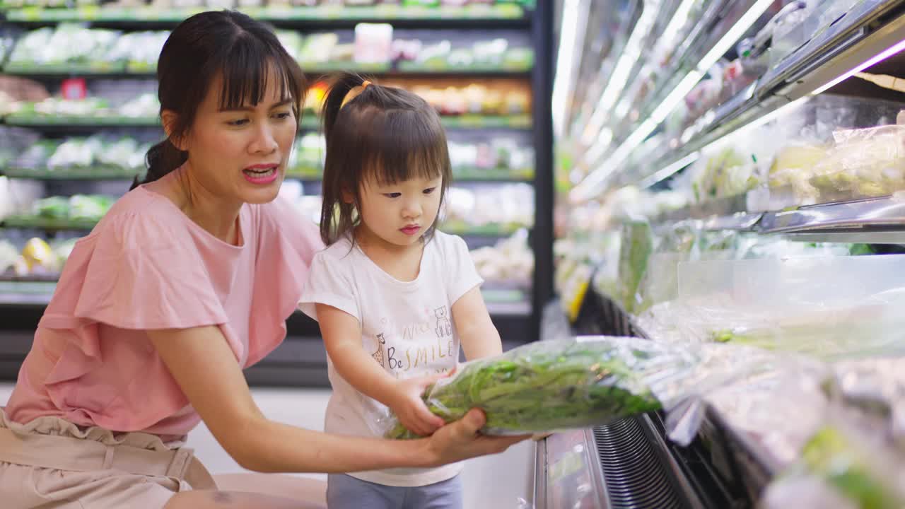 年轻的亚洲美丽的母亲拿着购物篮和她的孩子走在超市。她从货架上挑选绿色沙拉蔬菜，然后给她的小孩吃。健康的购物。视频素材