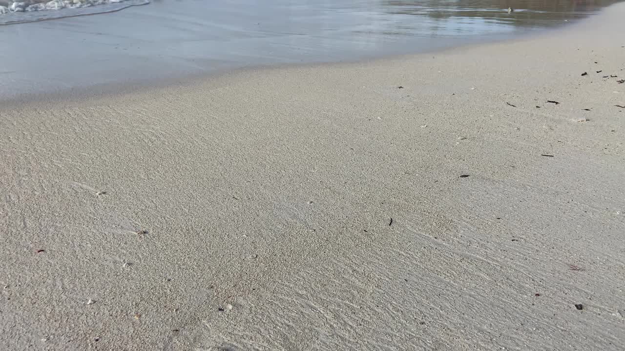 男性走在白色的沙滩上，海浪在他的脚下溅起水花。视频素材