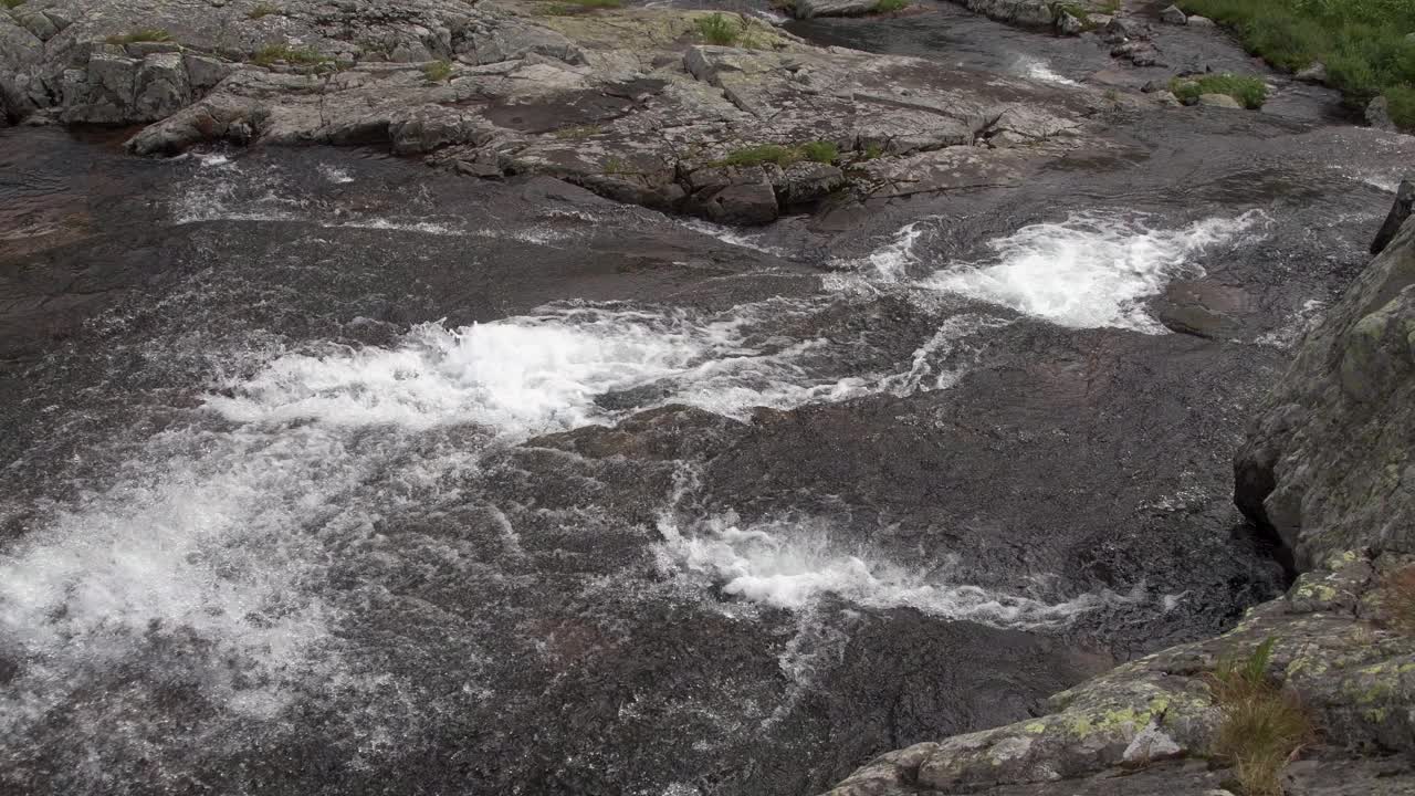 湍急的水流汩汩地沿着石径滴落。山河溅起泡沫，溅起水花。岩石海岸上的绿色植被。小溪奔流进山谷。视频素材