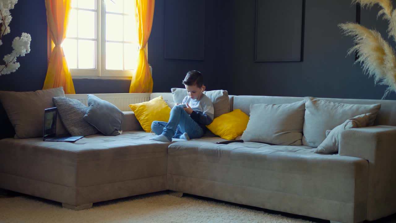 一个学龄前的男孩坐在家里的沙发上用手机和耳机进行在线教育。孩子使用在线学习应用程序。一个拿着小玩意儿的孩子。视频素材