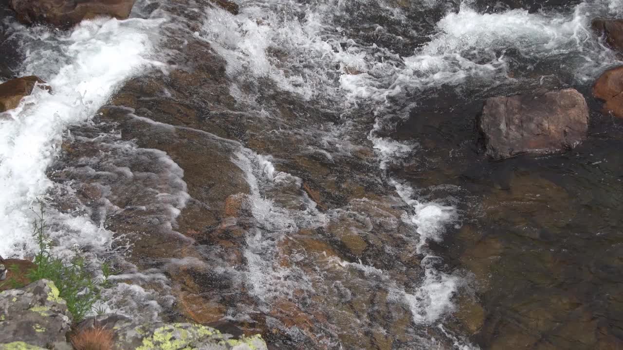 狂风暴雨的山涧从石床上泻下。河流沿着花岗岩墙流淌。水溅在岩石上形成水花。清澈小溪岸边的绿草。视频素材