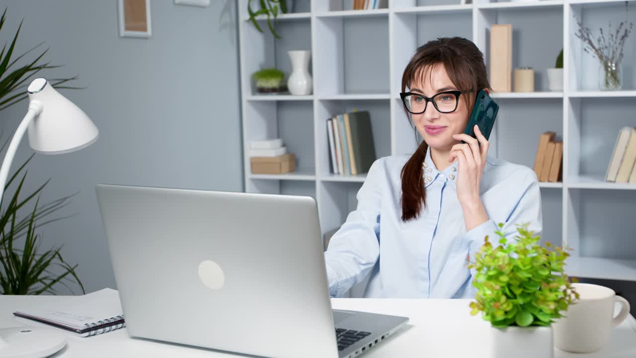 快乐的年轻商业女性用笔记本电脑打电话。快乐友好的女孩坐在时尚的办公室里通过智能手机说话。视频素材