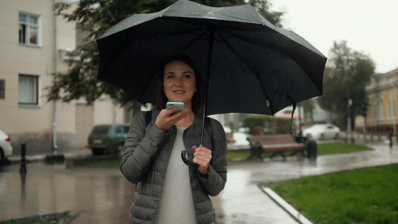 女孩用智能手机语音识别边走边撑着雨伞雨天雨天，口里口里念，晚上语音拨号信息，学生模特年轻上街头语音ai语音信息视频素材