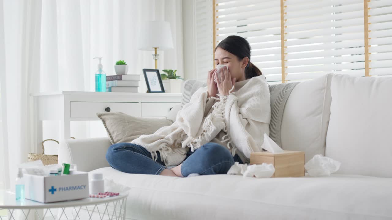 生病的年轻亚洲女人坐在沙发上的毯子下，用纸巾在家里打喷嚏。女性擤鼻涕，咳嗽或在家里用纸巾打喷嚏，患流感。寒热概念视频素材